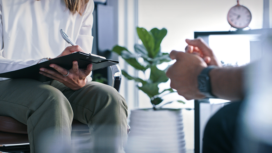 Close up of woman sitting on sofa writing on a clipboard, seated across from man; no faces visible
